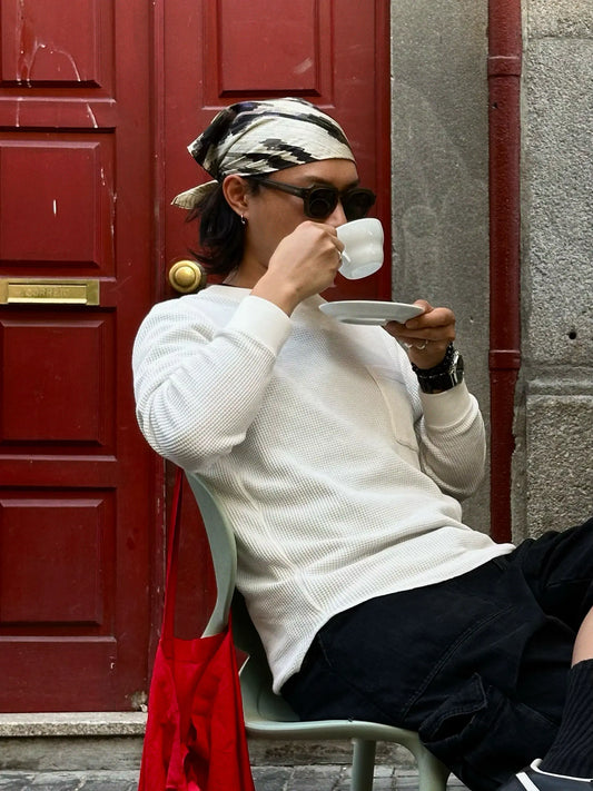 Man with dark grey and beige abstract leopard bandana in white top sitting on a chair holding a cup in front of a red door.