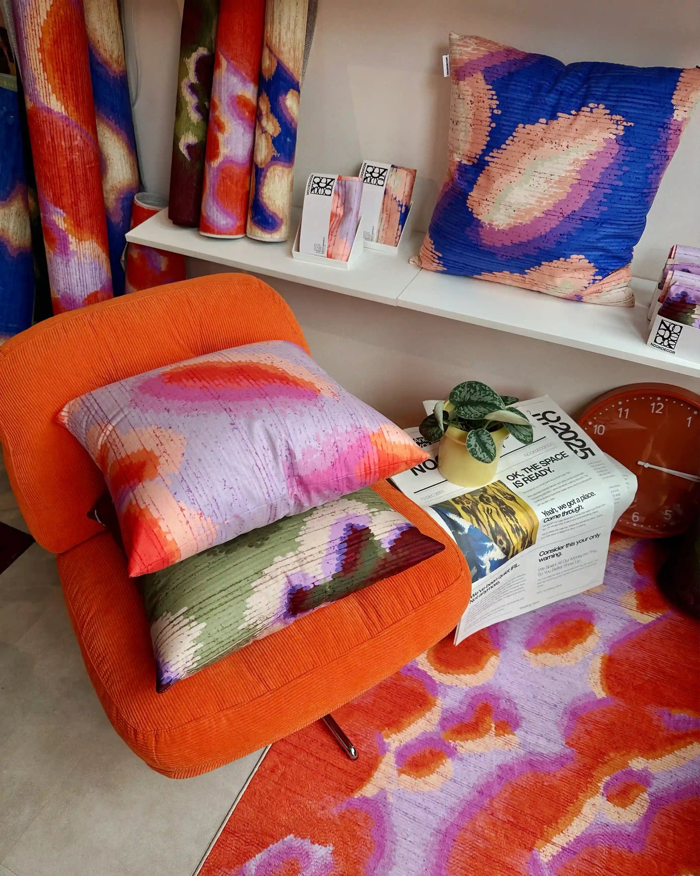 A 3x5' abstract camouflage area rug in orange red and pastel purple on the foreground. And three abstract graphic cushion covers layered on Ikea orange sofa and shelves, and rolled rugs placed at the corner in eclectic modern living room.