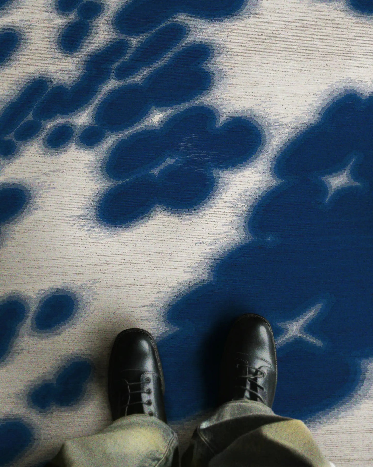 A man in trendy baggy jeans and black leather shoes stands on a faux wool, 3mm low pile height cowhide rug featuring abstract floral patterns in Klein blue and off white hues, radiating a contemporary vibe in a stylish setting.