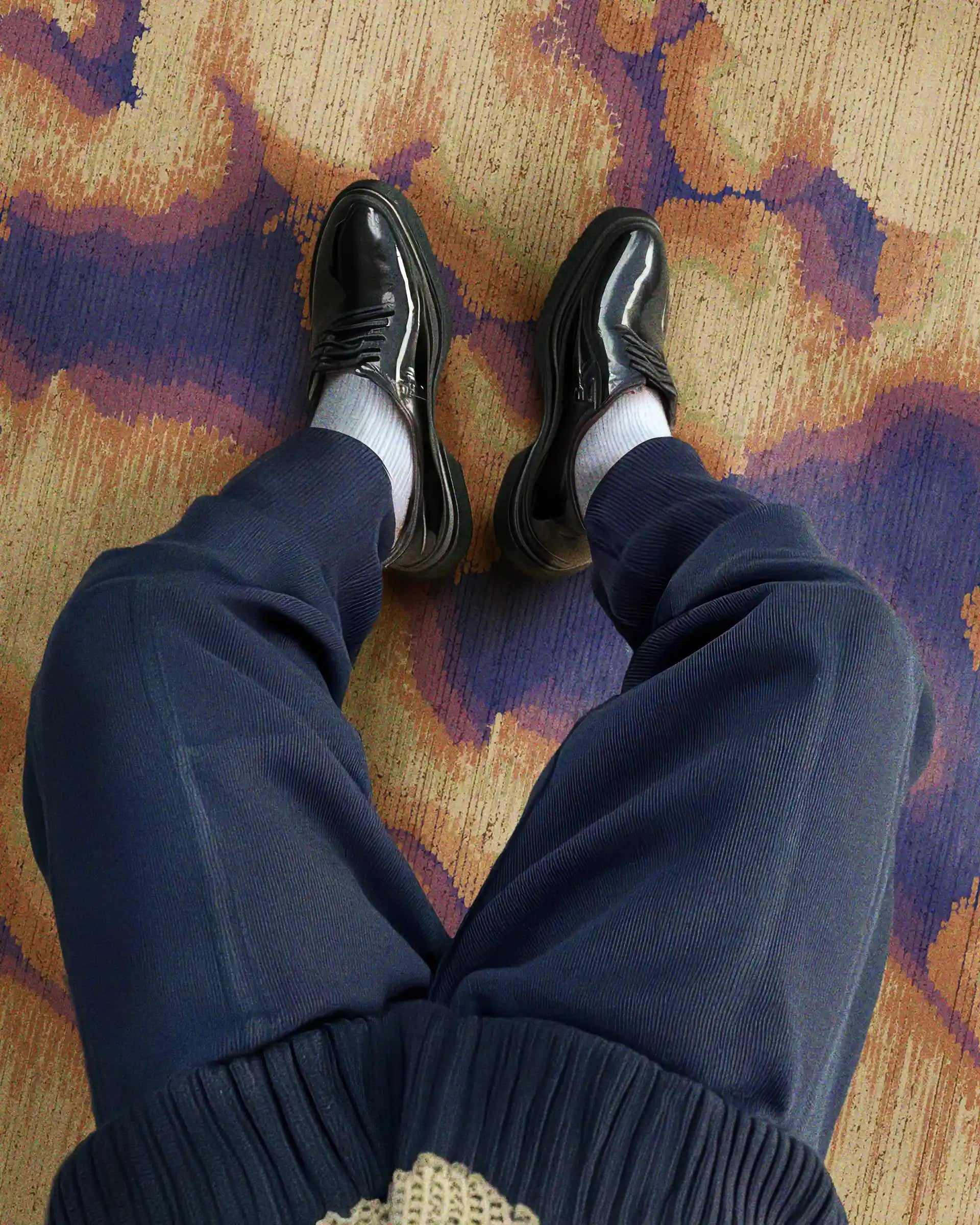 Man wearing black leather shoes and navy pants resting his legs on an eclectic home decor, the abstract purplish-blue and creamy yellow camouflage area rug in his modern dopamine living room.