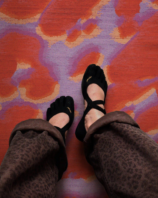 Woman wearing Vibram FiveFingers training shoes and leopard pants resting her legs on an eclectic home decor, the abstract orange, red and pastel purple camouflage area rug in the modern dopamine living room.