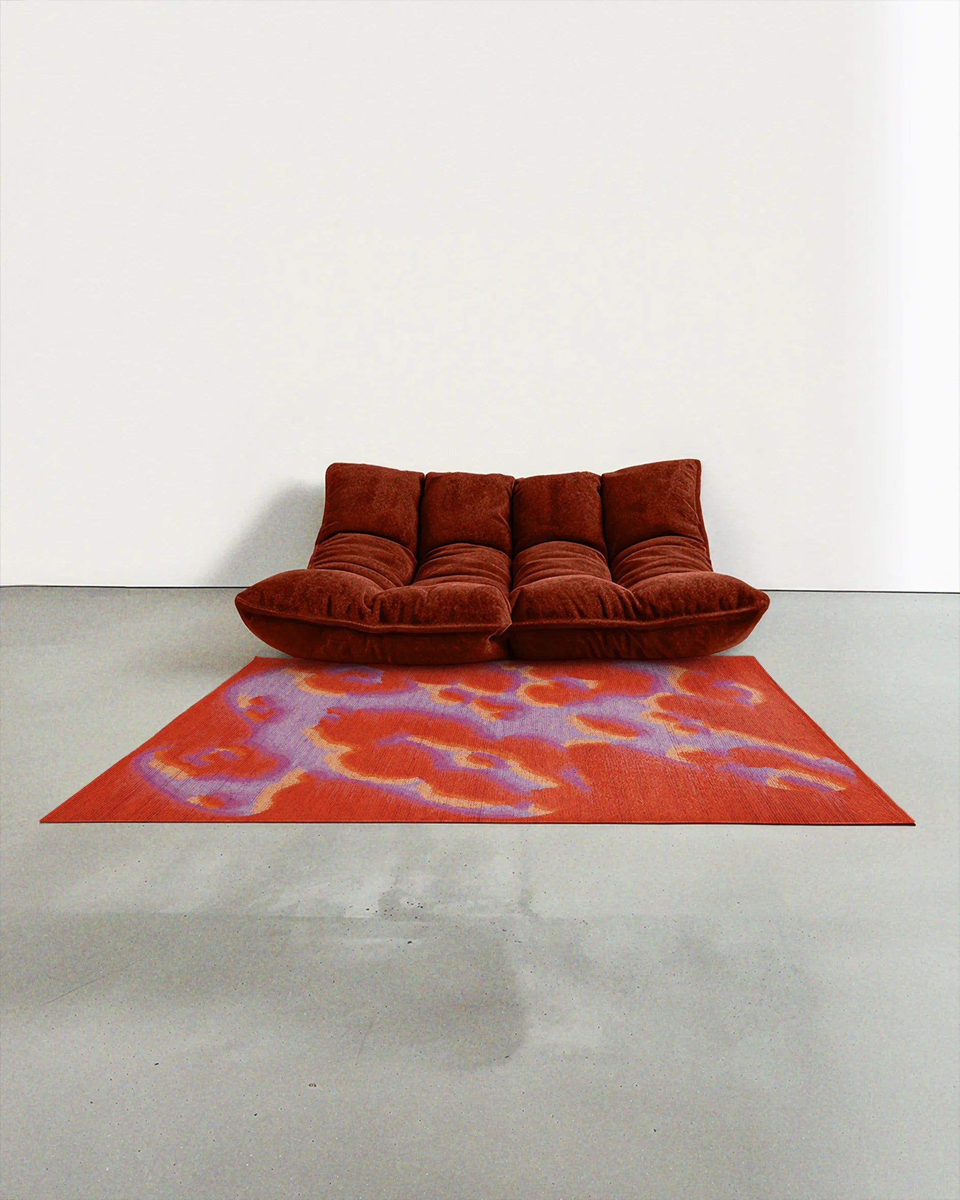 A dopamine abstract camouflage area rug measuring 5ft by 7ft, in orange, red, and pastel purple tones, placed in a contemporary modern studio with a dark rusty orange two-seat sofa.