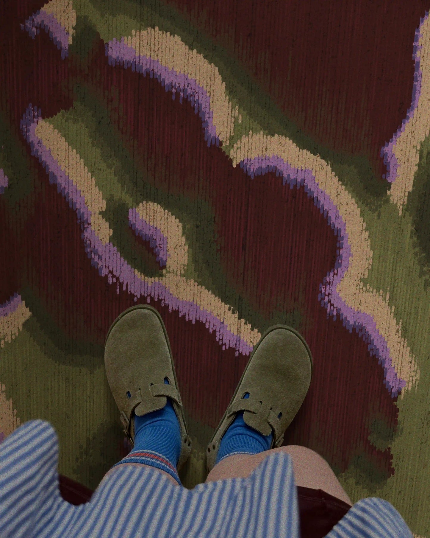 A woman standing on a dopamine abstract camouflage area rug in burgundy and love green hues, wearing olive green sandals, blue socks, and a blue-and-white-striped shirt, in her eclectic fall-vibe living room, in a maximalist apartment.