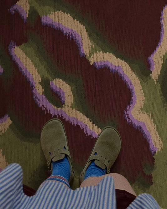 A woman standing on a dopamine abstract camouflage area rug in burgundy and love green hues, wearing olive green sandals, blue socks, and a blue-and-white-striped shirt, in her eclectic fall-vibe living room, in a maximalist apartment.
