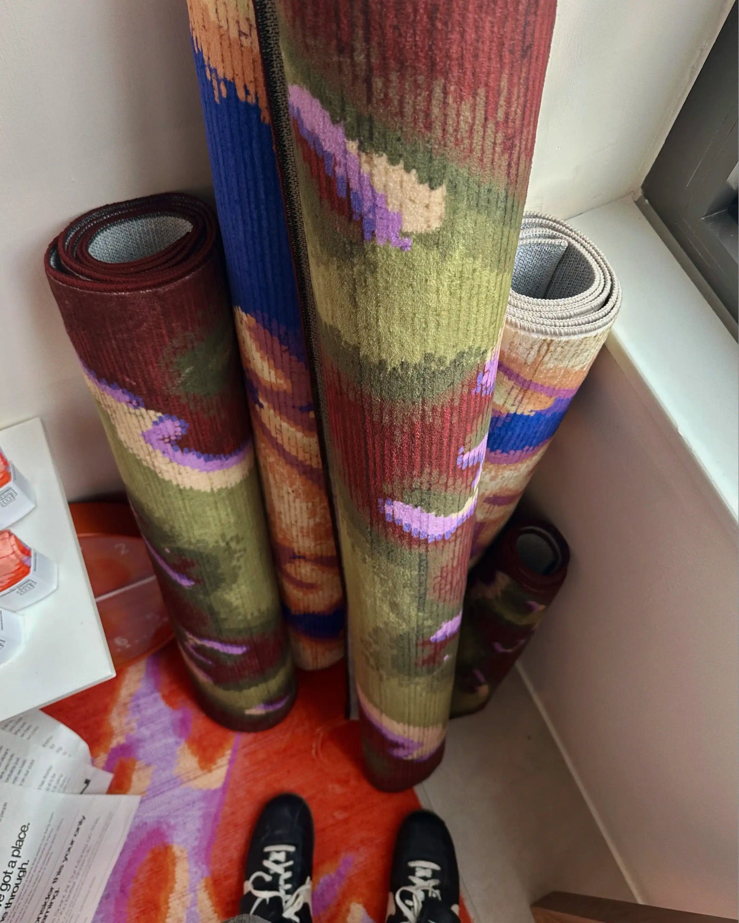 A man in the NOOKDECOR colorful showroom is standing on an abstract camouflage of orange, red, and purple hues. In contrast, the purple and creamy beige rug and the burgundy and olive green rug are being rolled up against the plain wall.