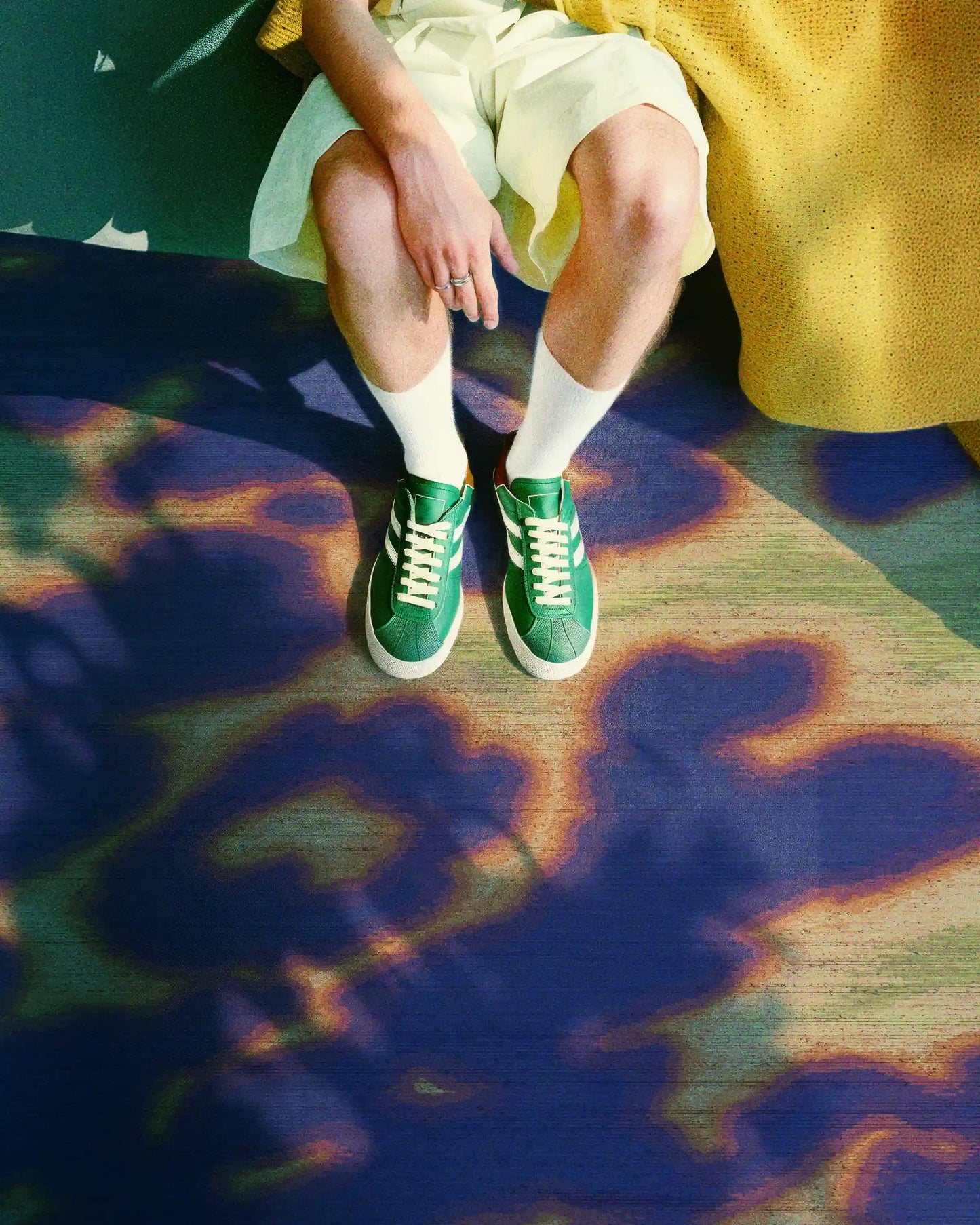 Man sitting on sofa wearing trendy outfit with light yellow shorts, white socks and green sneakers, resting his legs on an abstract cowhide area rug in purplish blue, light green and orange hues. Dopamine home decor for an eclectic living room.