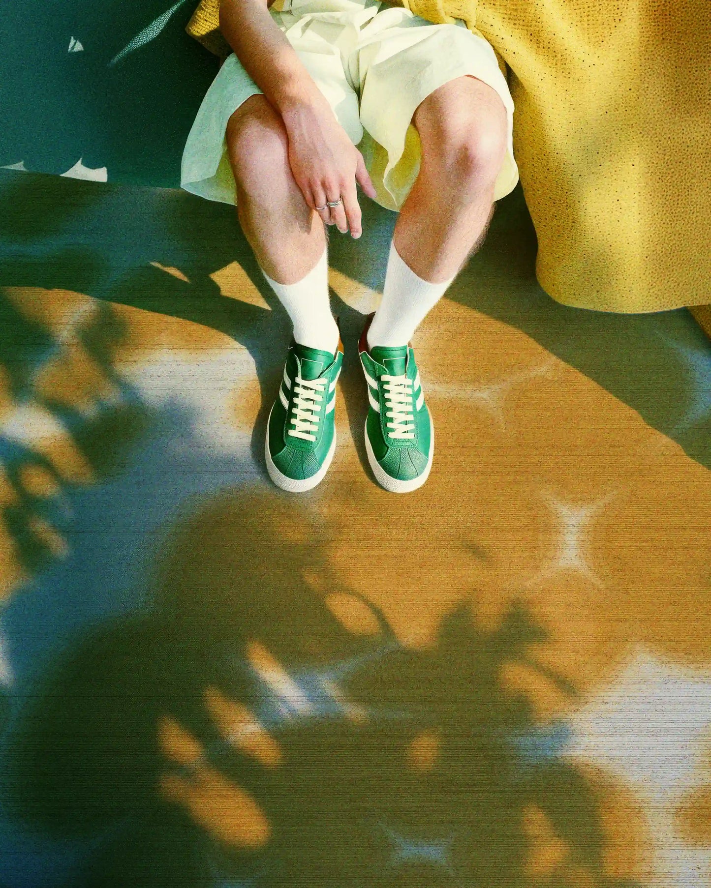 A man resting on a floral cowhide area rug in yellow and off white hues wearing light yellow shorts, white socks, and a pair of green sneakers in a sunlit living room.