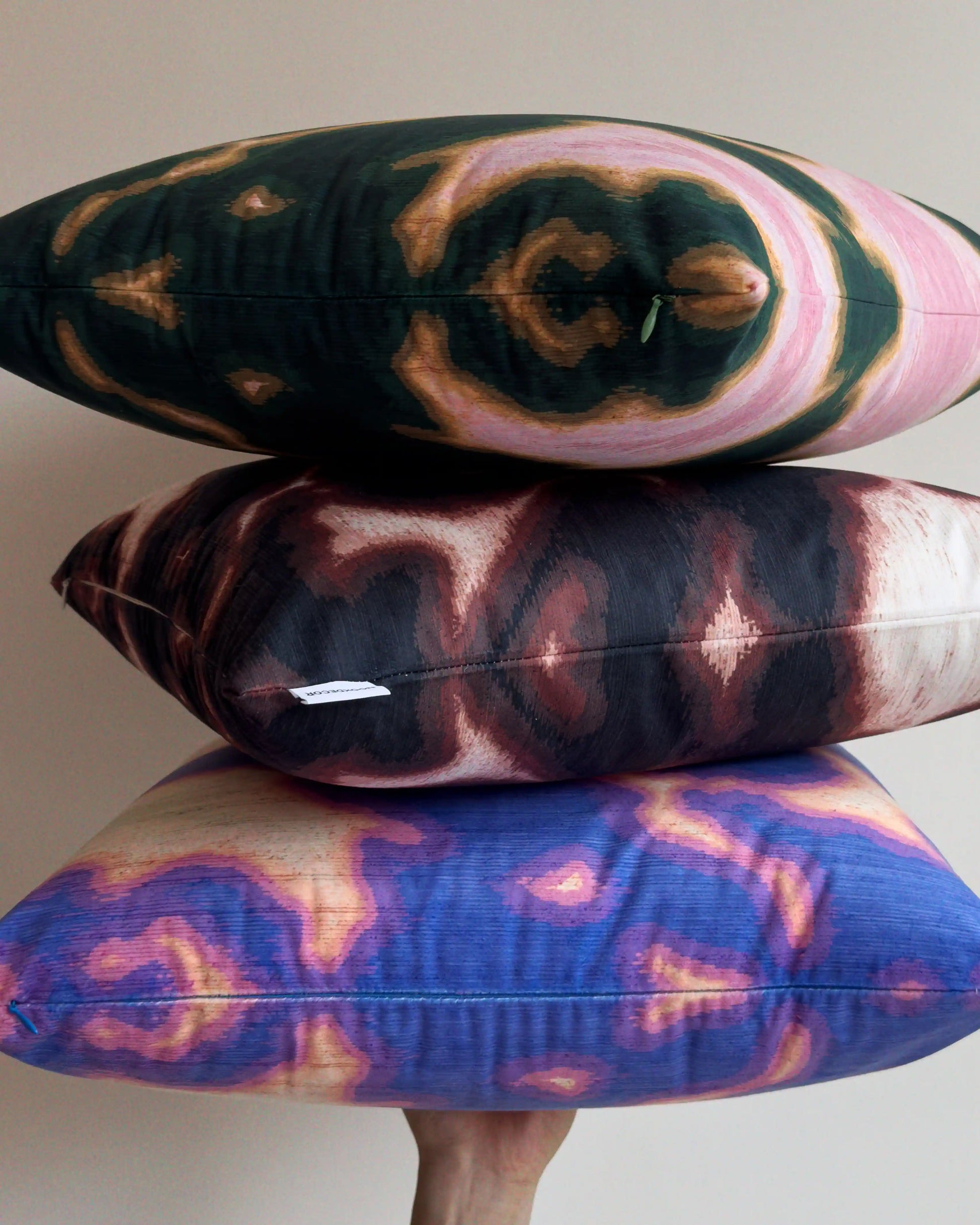 Close up of set of 3 coordinating throw pillows in dopamine colorful cushion covers held against beige white wall, showing mix of abstract floral cowhide for bold and eclectic home decor styling. Including, Pink and dark green/ Purplish blue and light green and dark brown and off white.