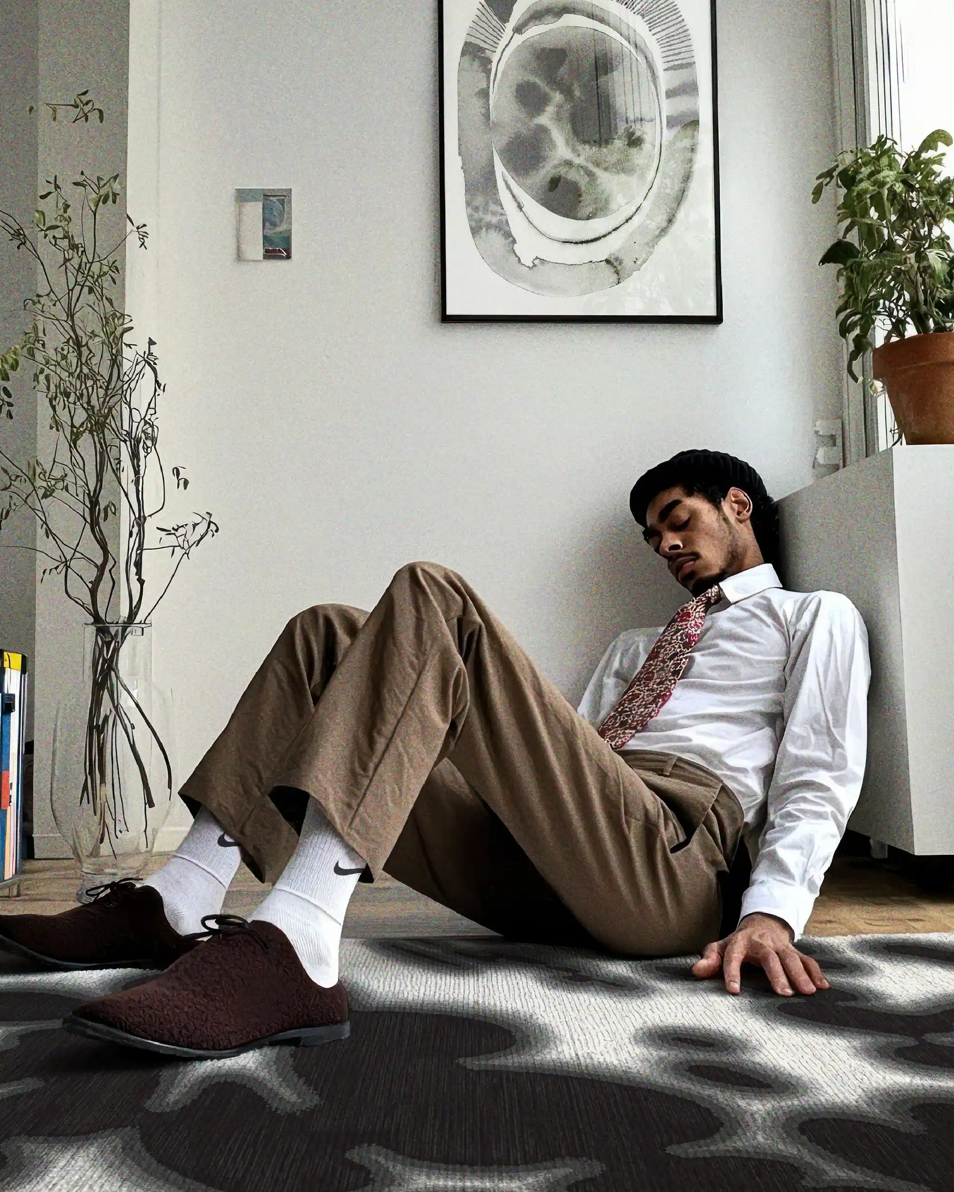 A man in a trendy casual suit, white shirt, and brown trousers rests on the floor of a black and off white floral cowhide area rug with abstract artwork painted by Neil Wang, the founder of NOOKDECOR, in a minimalist Scandinavian living room.