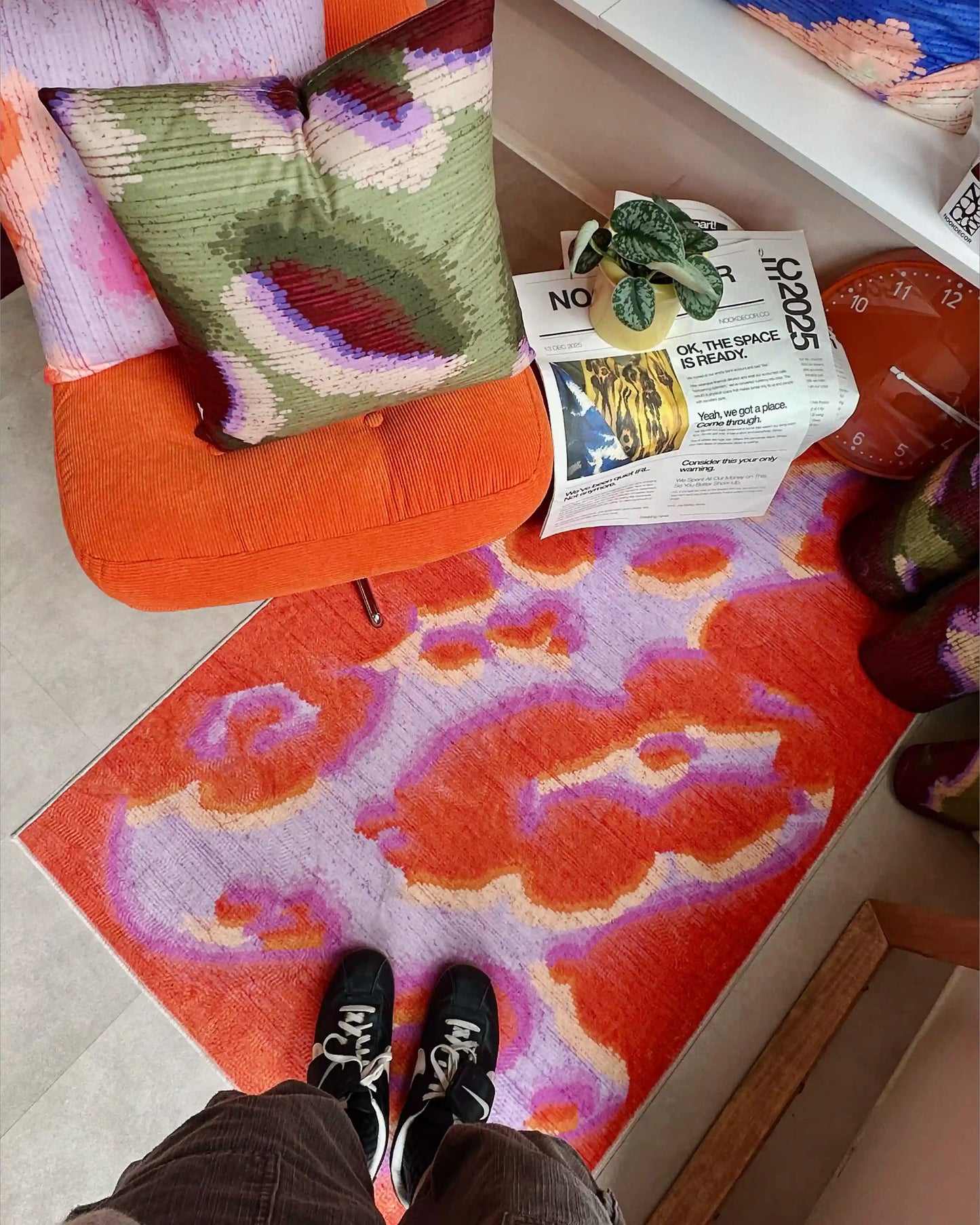 Man standing on abstract camouflage area rug in orange red and pastel purple, And 2 abstract graphic cushion covers layered on ikea orange sofa in modern living room.