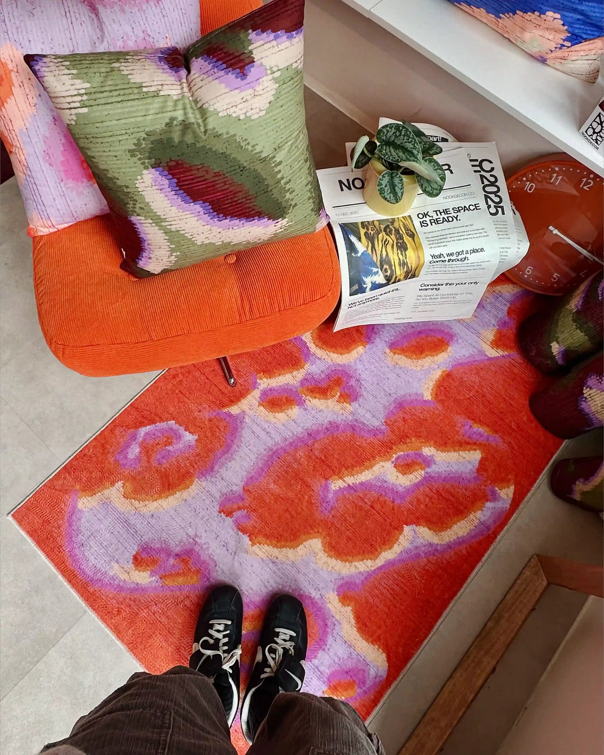 Man standing on abstract camouflage area rug in orange red and pastel purple, And 2 abstract graphic cushion covers layered on ikea orange sofa in modern living room.