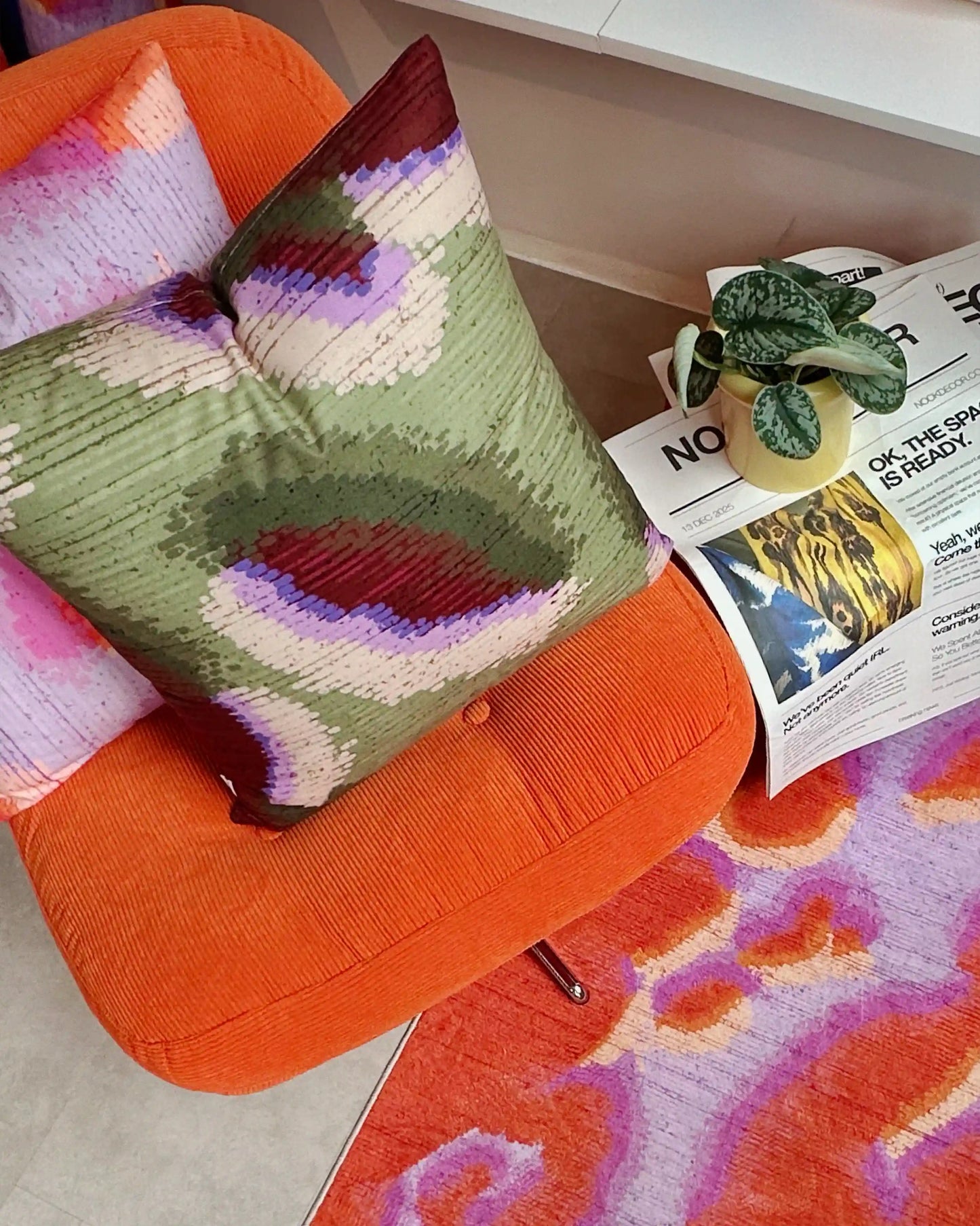 A 3x5' abstract camouflage area rug in orange red and pastel purple on the foreground. And 2 abstract graphic cushion covers layered on Ikea orange sofa, and a house plant on newspaper in eclectic modern living room.