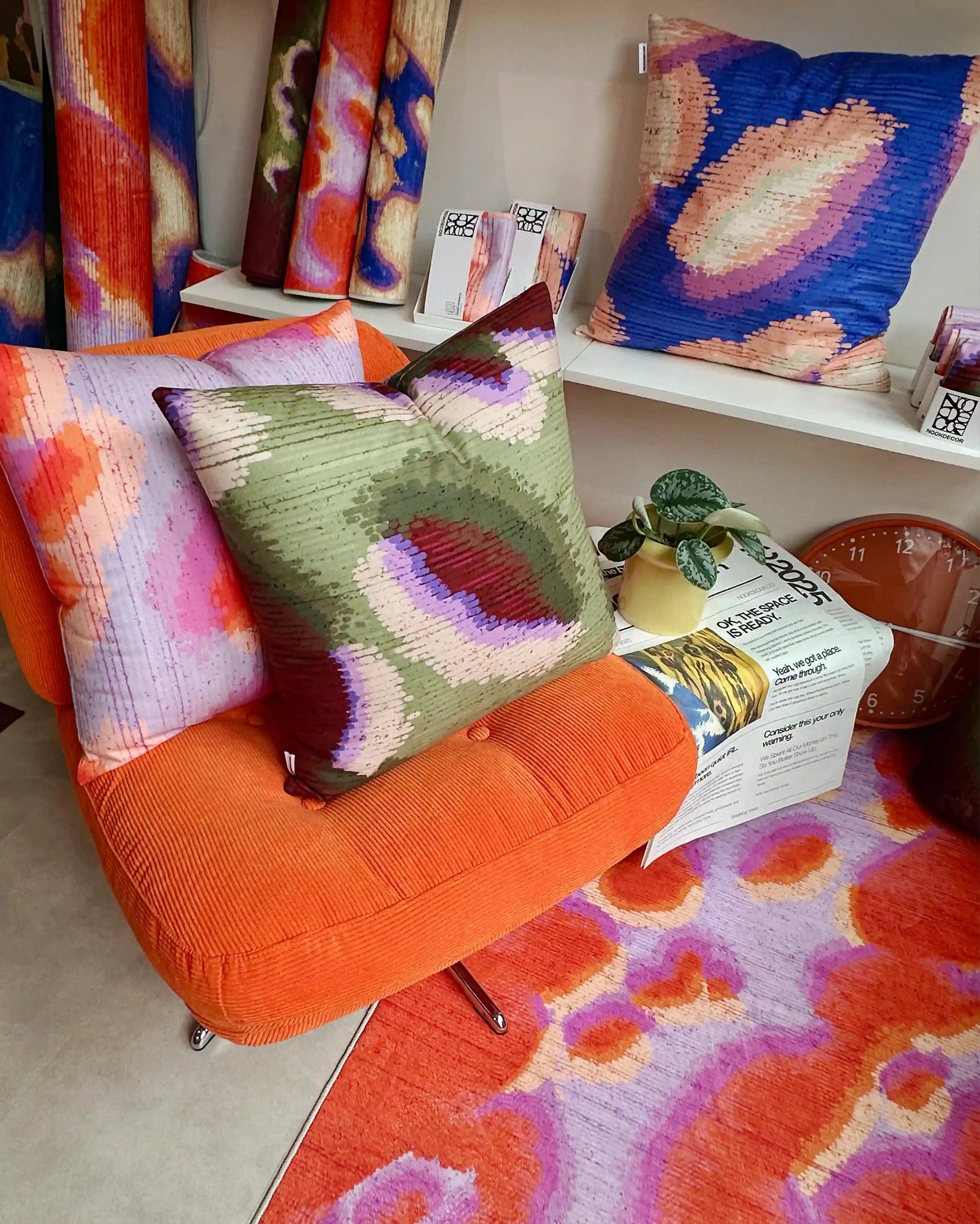 Abstract camouflage area rug in orange red and pastel purple on the foreground. And three abstract graphic cushion covers layered on Ikea orange sofa and shelves, and rolled rugs placed at the corner in modern living room.