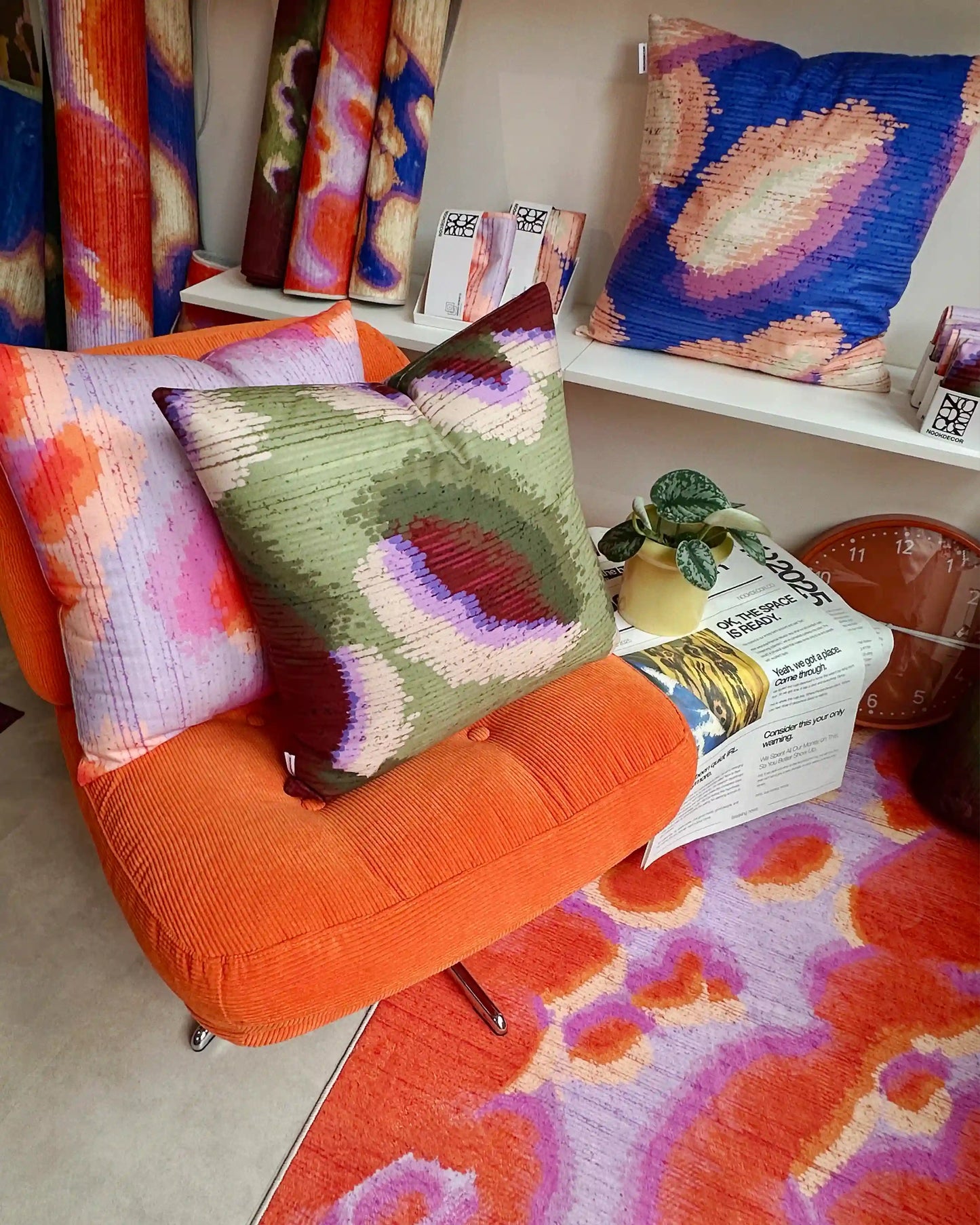 Abstract camouflage area rug in orange red and pastel purple on the foreground. And three abstract graphic cushion covers layered on Ikea orange sofa and shelves, and rolled rugs placed at the corner in modern living room.