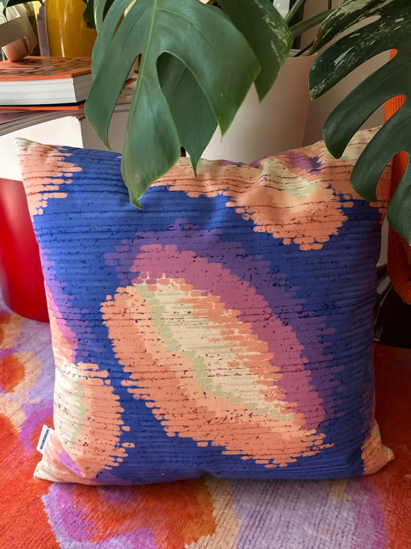 Colorful purplish-blue and coral-beige dopamine cushion cover placed on a red-and-purple rug from NOOKDECOR, with a houseplant and a stool next to it in an eclectic maximalist studio.