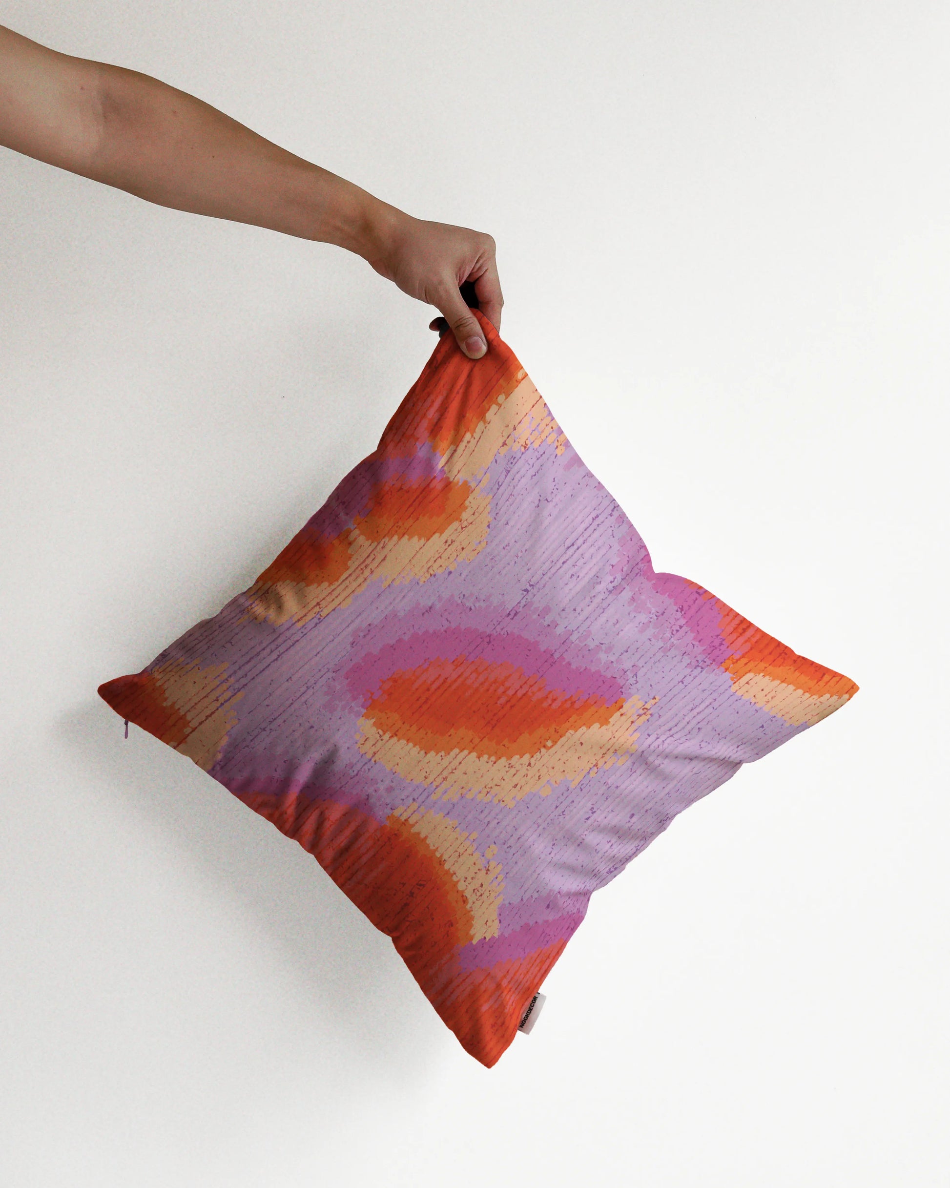 A hand holding a fall vibe, abstract camouflage cushion cover in orange red, pastel purple hues, against a minimalist beige background, perfect for eclectic dopamine decor in a modern apartment.