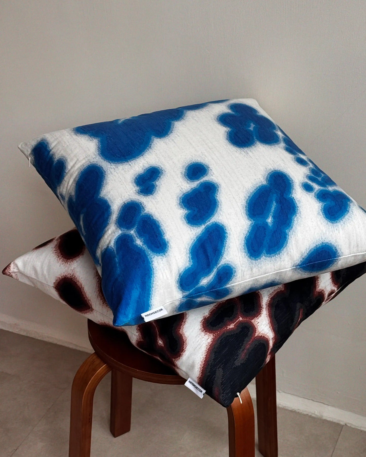 Set of two patterned pillows on a wooden stool against a plain wall. Features Klein blue and off white and dark brown and off white abstract floral cowhide pattern.