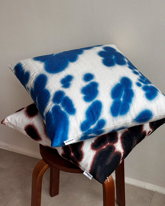 Set of two patterned pillows on a wooden stool against a plain wall. Features Klein blue and off white and dark brown and off white abstract floral cowhide pattern.