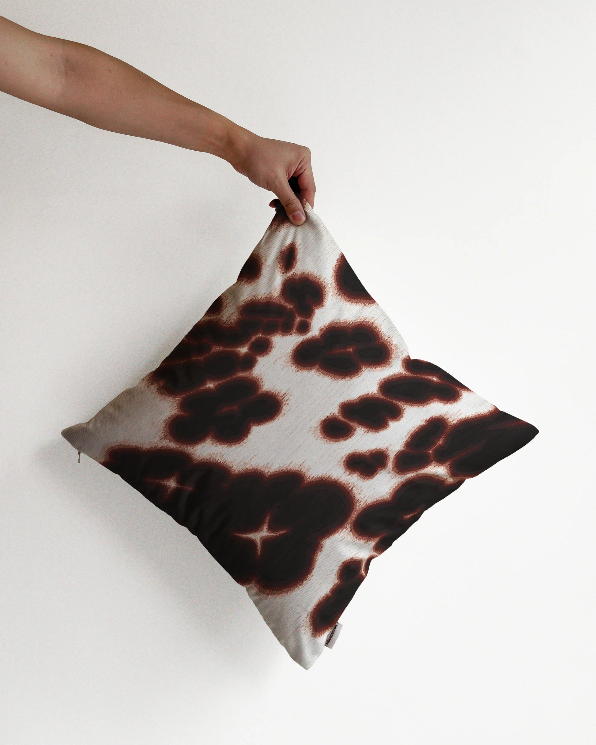 A hand holding a bold, abstract floral and cowhide cushion cover in brown and off white hues, against a minimalist beige background, perfect for eclectic dopamine decor in a modern apartment.