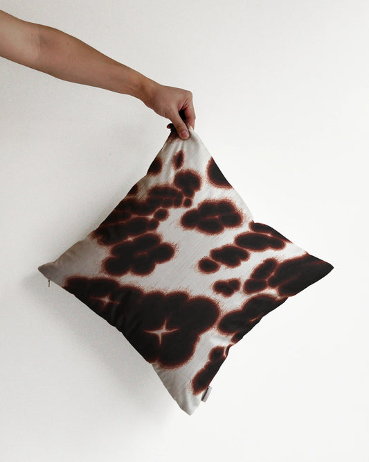 A hand holding a bold, abstract floral and cowhide cushion cover in brown and off white hues, against a minimalist beige background, perfect for eclectic dopamine decor in a modern apartment.