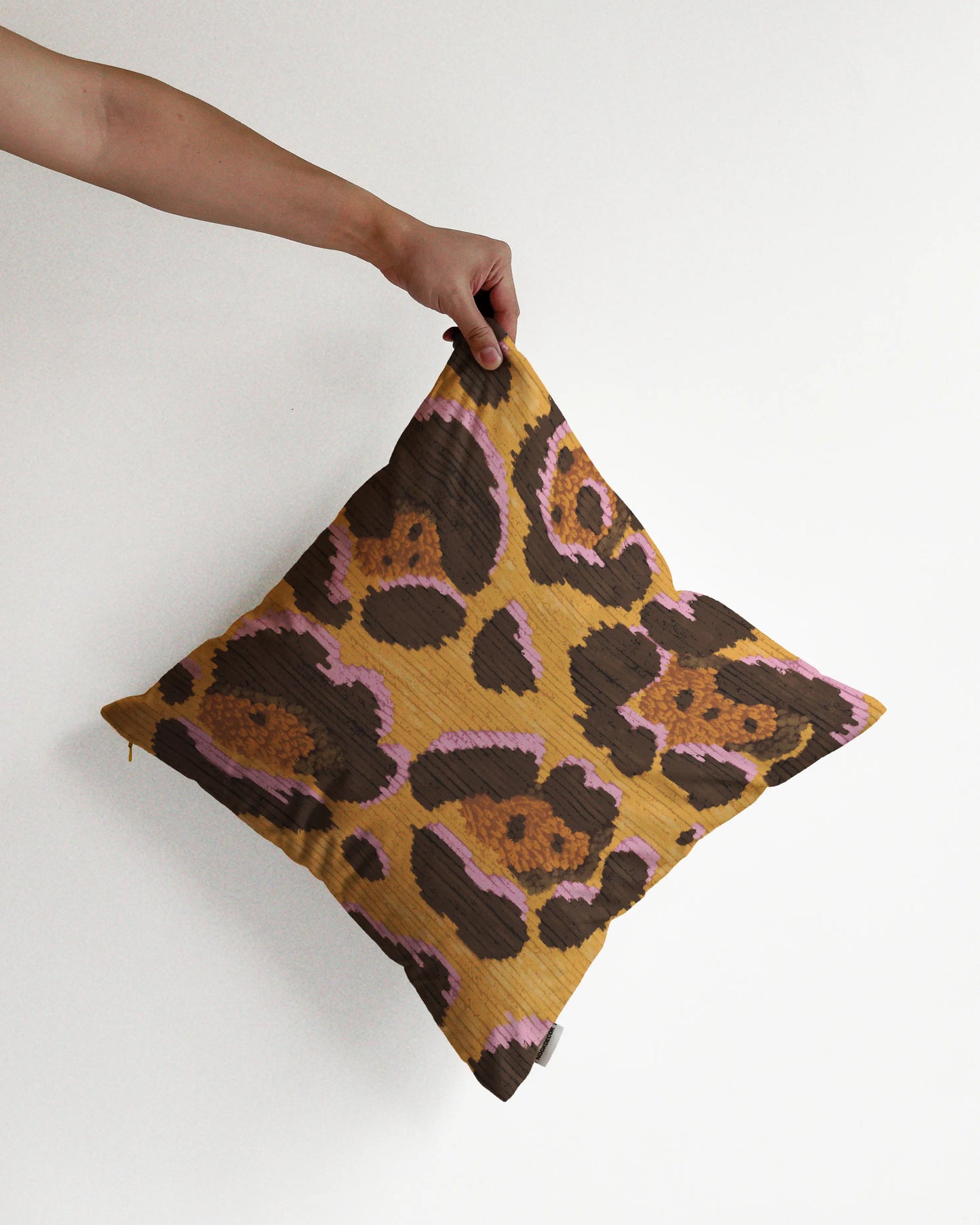 A hand holding a bold abstract floral and leopard cushion cover in yellow, pink and brown hues, against a minimalist beige background, perfect for eclectic dopamine decor in a maximalist modern apartment.