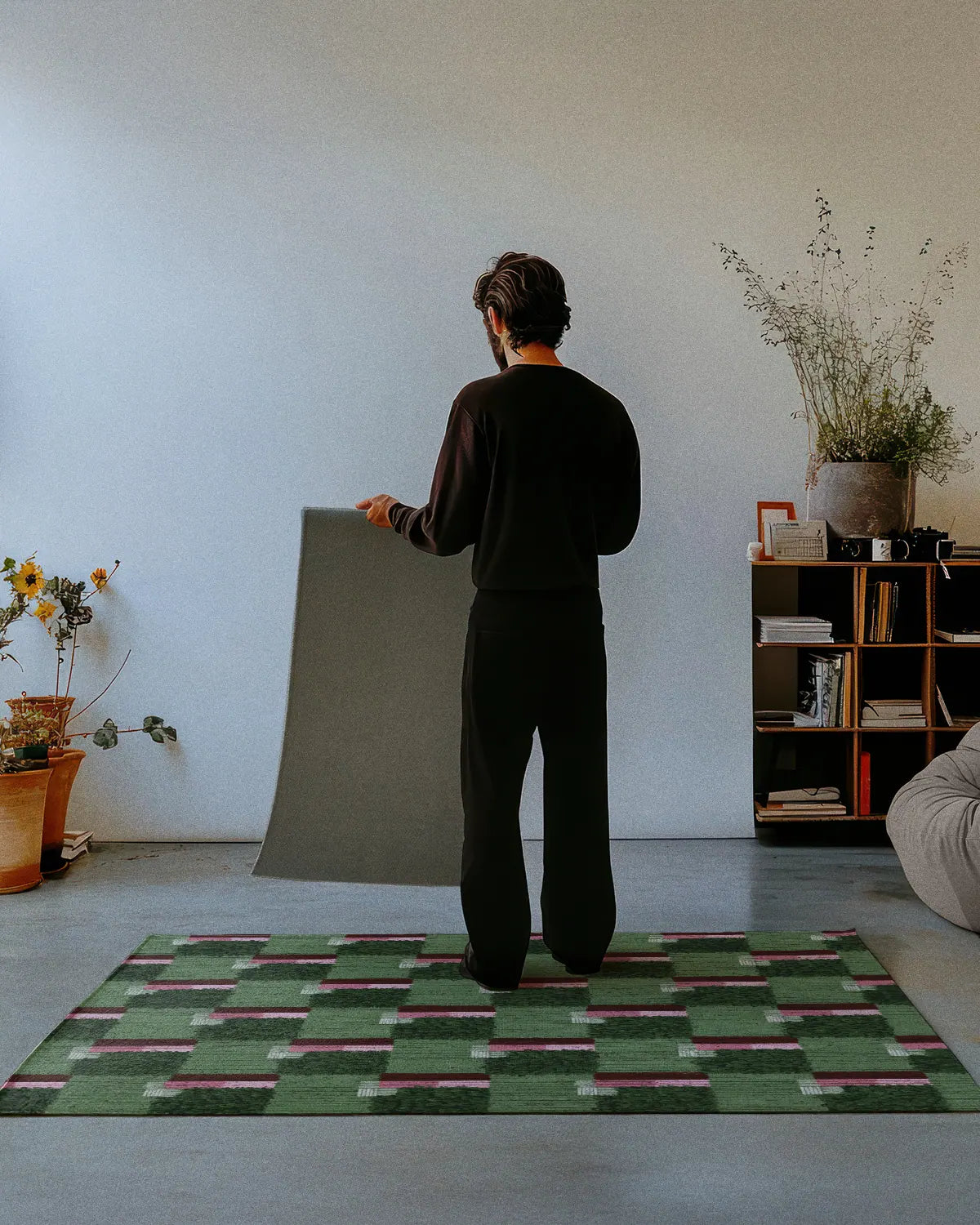 A man dressed in all black stands on a sage green checkered rug 4x6, showing the nonskid rug backing of a 2x3 rug in a modern studio.