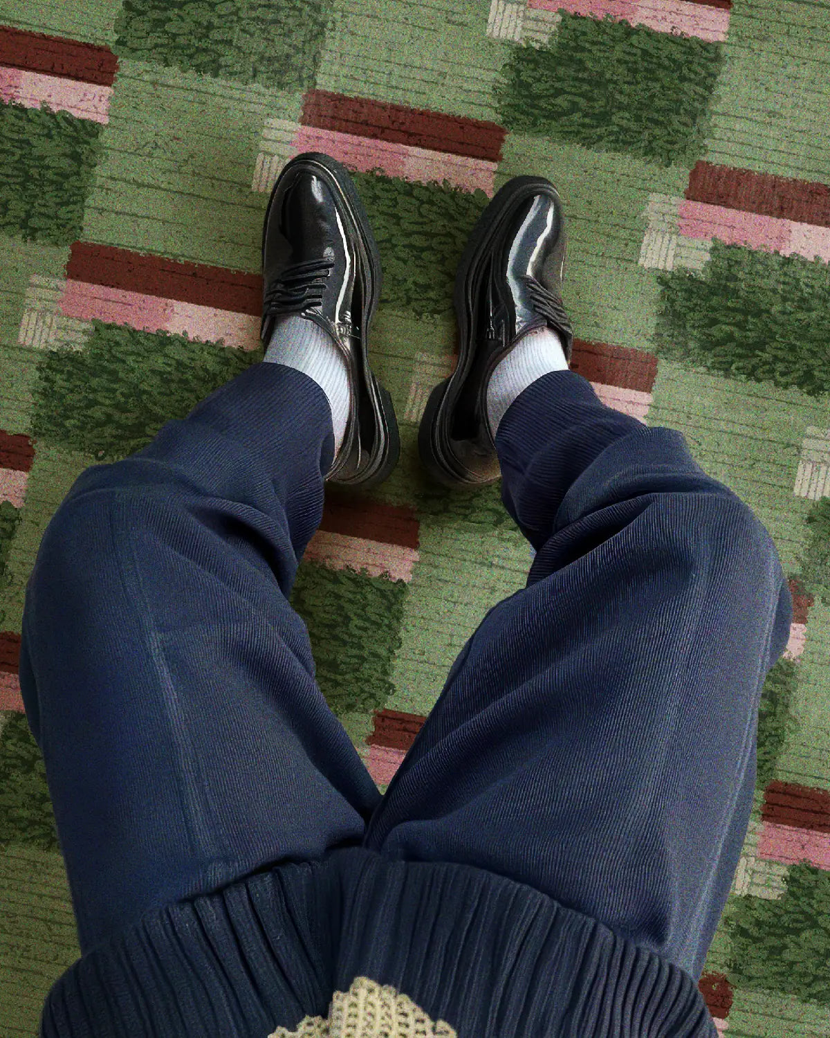 A man wearing navy sweat pants and black leather shoes is relaxing on a sage green checkered rug 8x10 with a slight touch of burgundy and pink hues in a modern living room. 