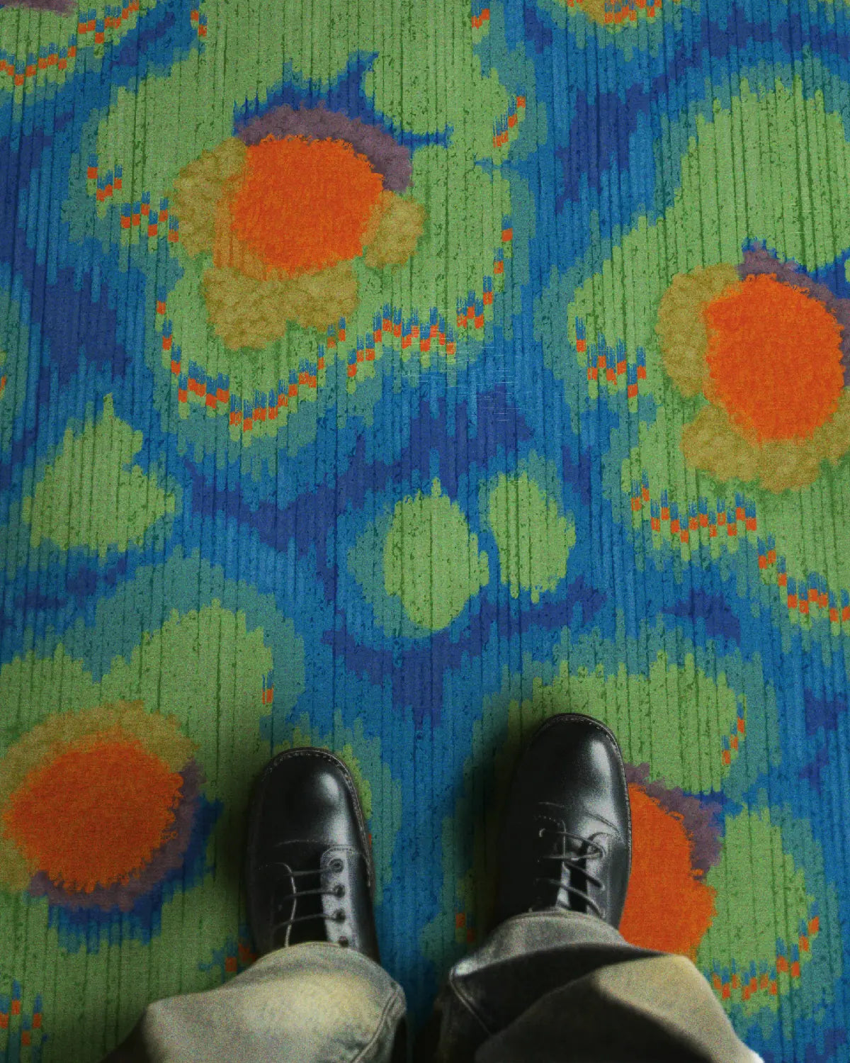 A man wearing baggy jeans and black leather shoes stands on an eclectic bright colored area rug features cute floral patterns in Klein blue, green and orange hues.