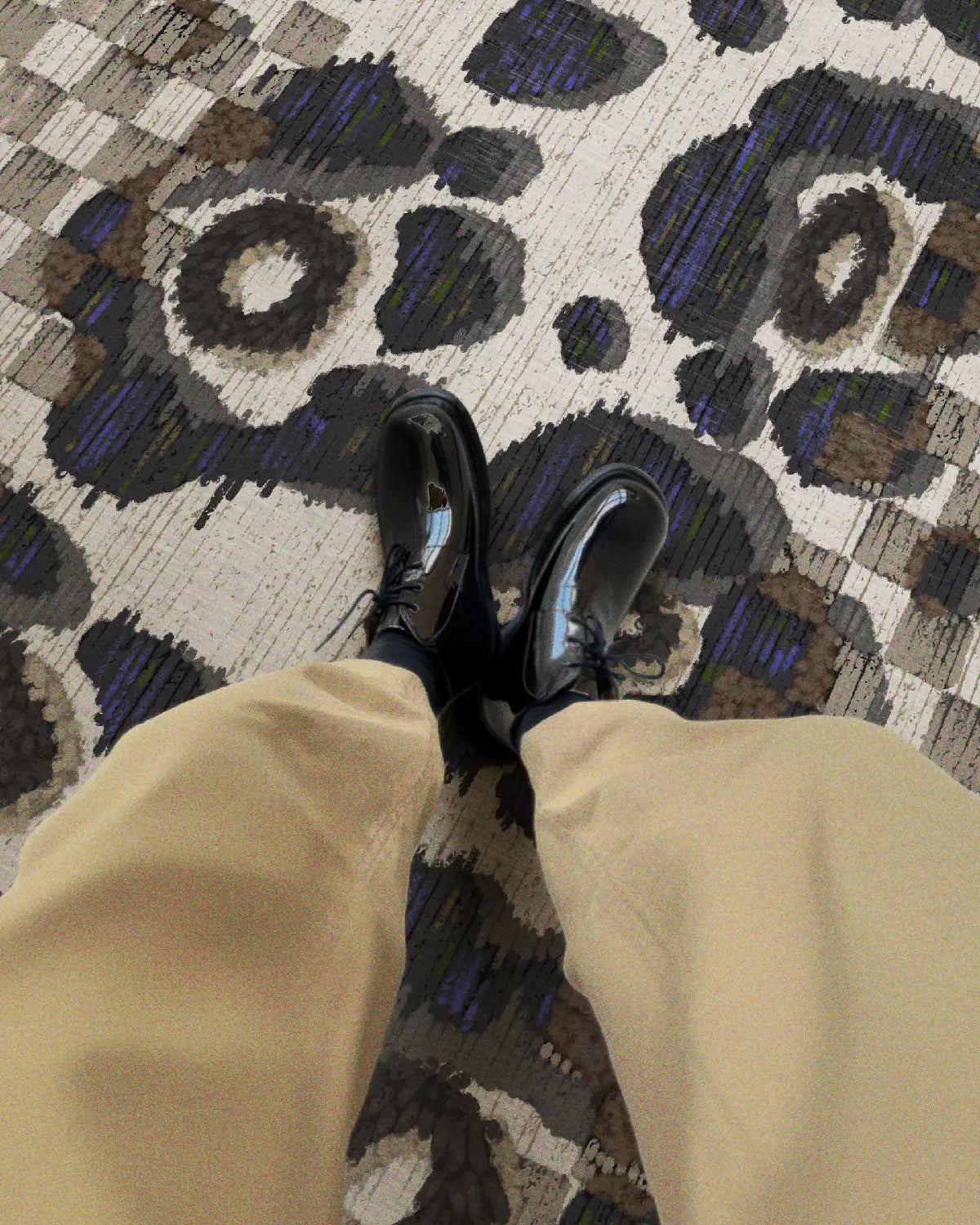 A man relaxing his legs on the black and white floral checkered area rug and wearing khaki pants and black leather shoes in a mid century modern living room.