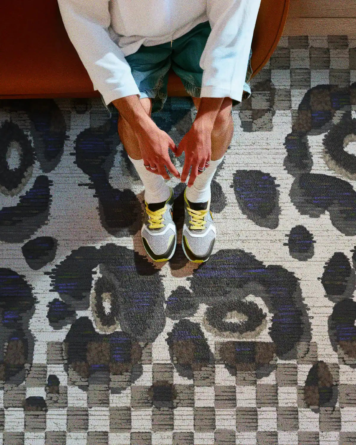 A man is sitting on a rust sofa, relaxing on a black and white checkered rug with abstract floral patterns, and wearing a white tee, demine shorts, and trendy sneakers.