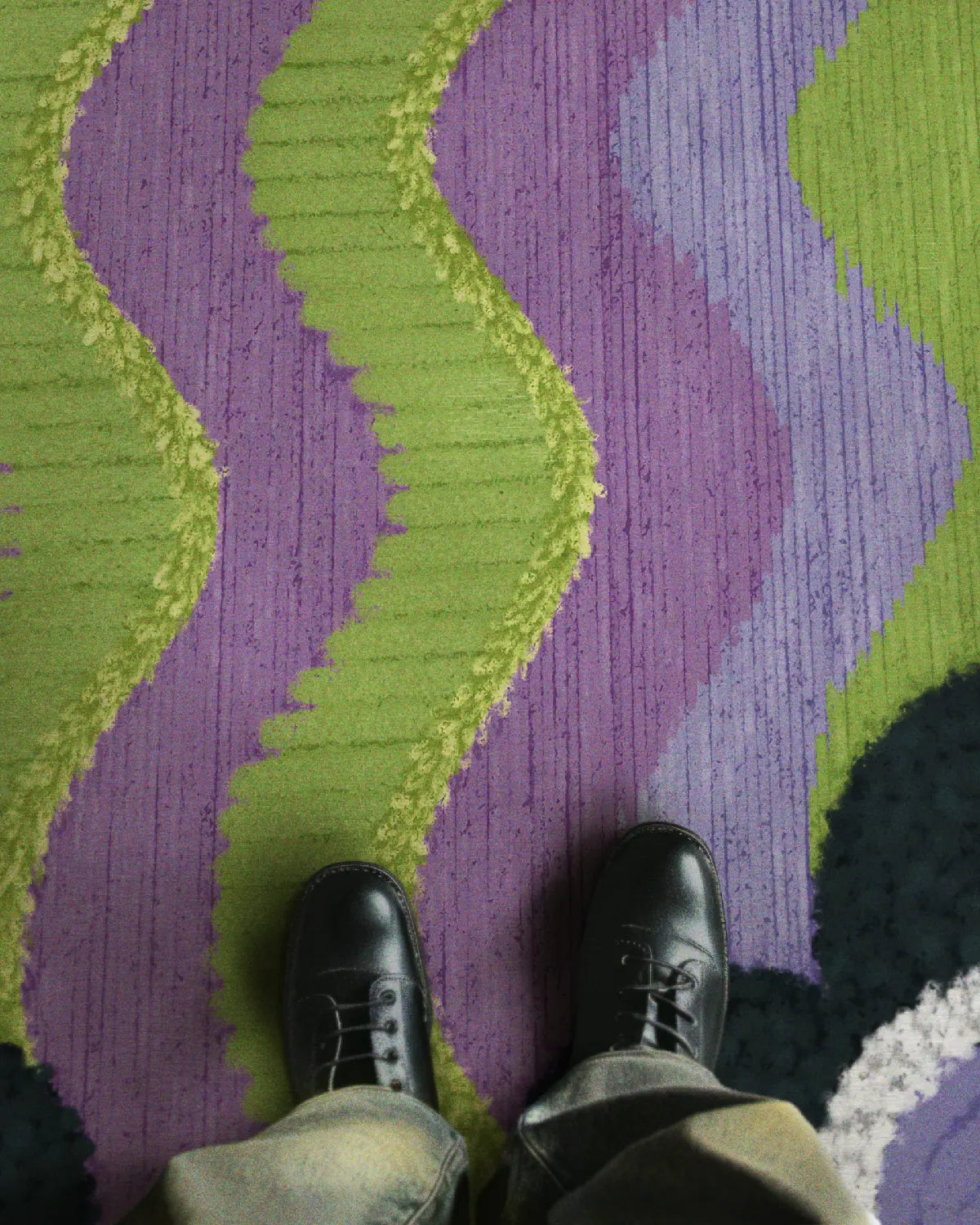 A man stands on the wavy patterned purple and green geometric rug, wearing faded baggy jeans and black leather shoes in a modern home.