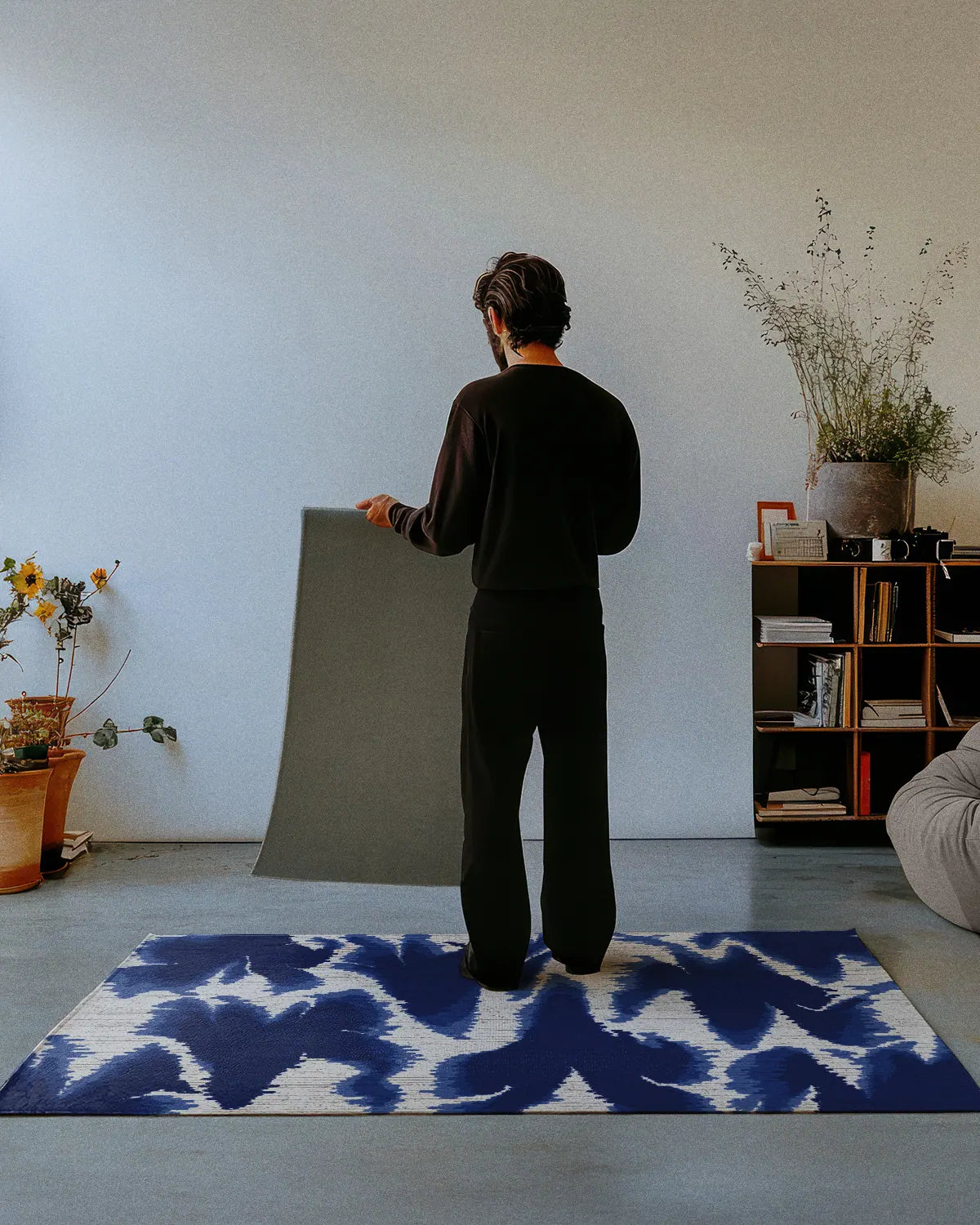 A man dressed in all black stands on a mid century modern area rug 4x6 in aura design blue and white hues, showing the non skid rug backing of a 2x3 rug in a studio.