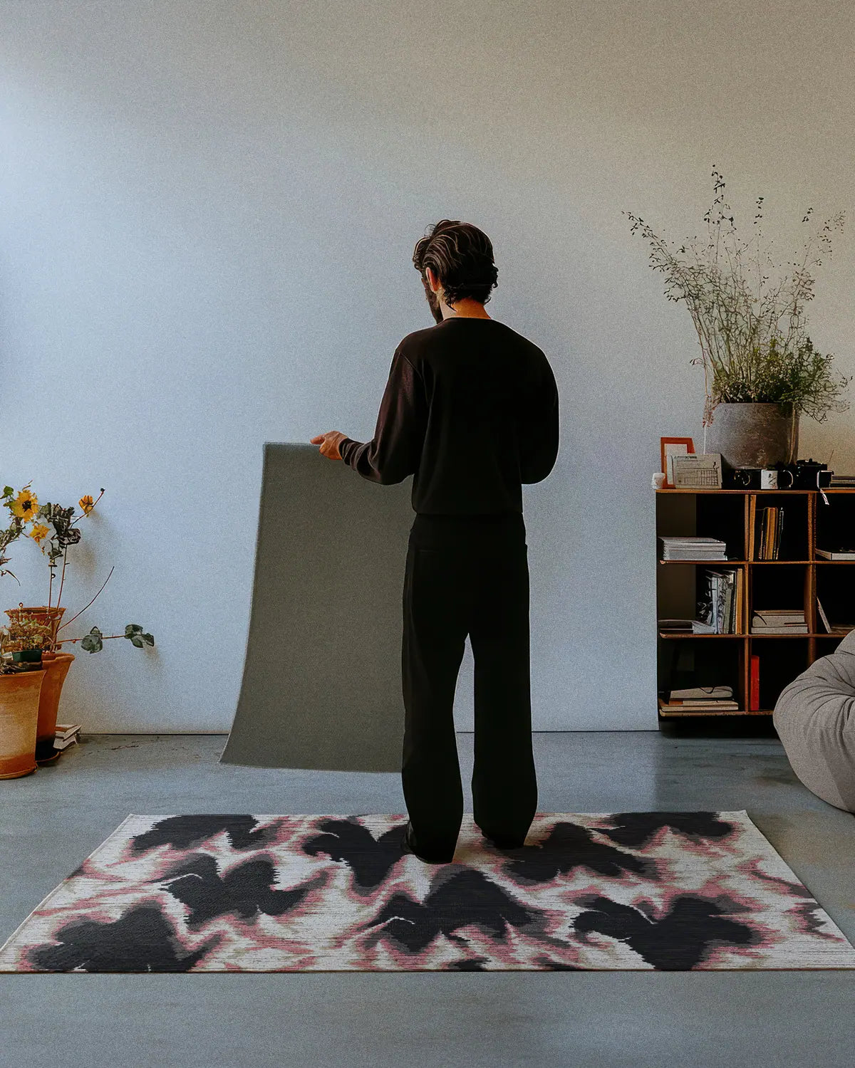 A man dressed in all black stands on a contemporary area rug 4x6, aura design in pink and grey hues, showing the non skid rug backing of a 2x3 rug in an eclectic style interior studio.
