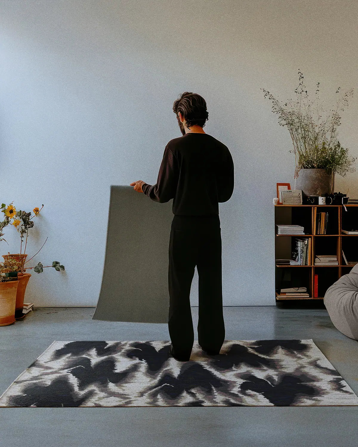 A man dressed in all black stands on a Scandinavian rug 4x6 in aura design grey, black and white hues, showing the non skid rug backing of a 2x3 rug in a mid century modern studio.
