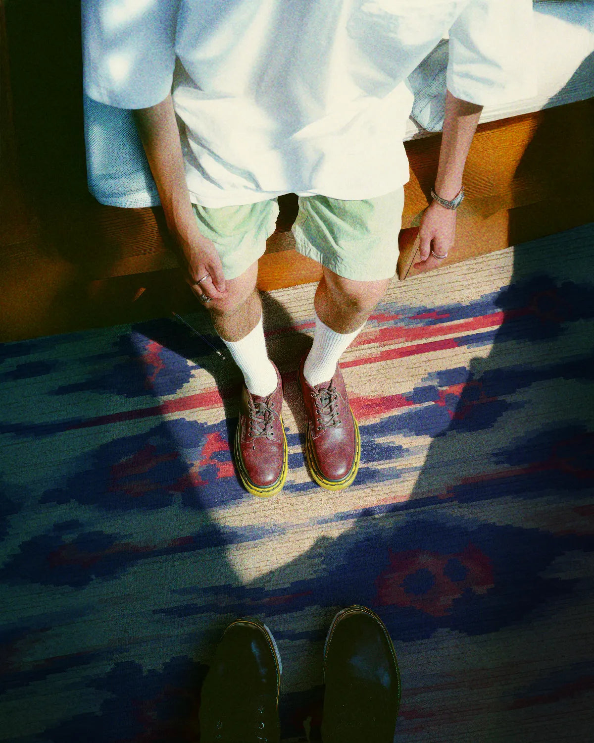 A man stands on an eclectic beige, blue, and coral colored area rug, wearing a trendy summer outfit in his modern living room.