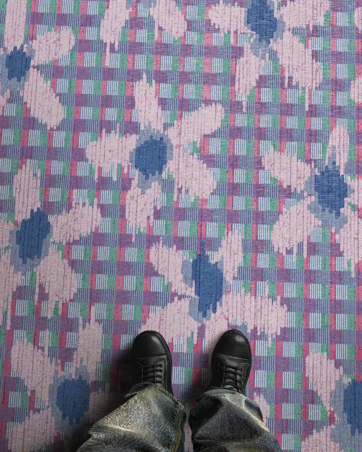 A man wearing trendy baggy jeans and black leather shoes stands on a pastel purple floral checkered rug in his living room.