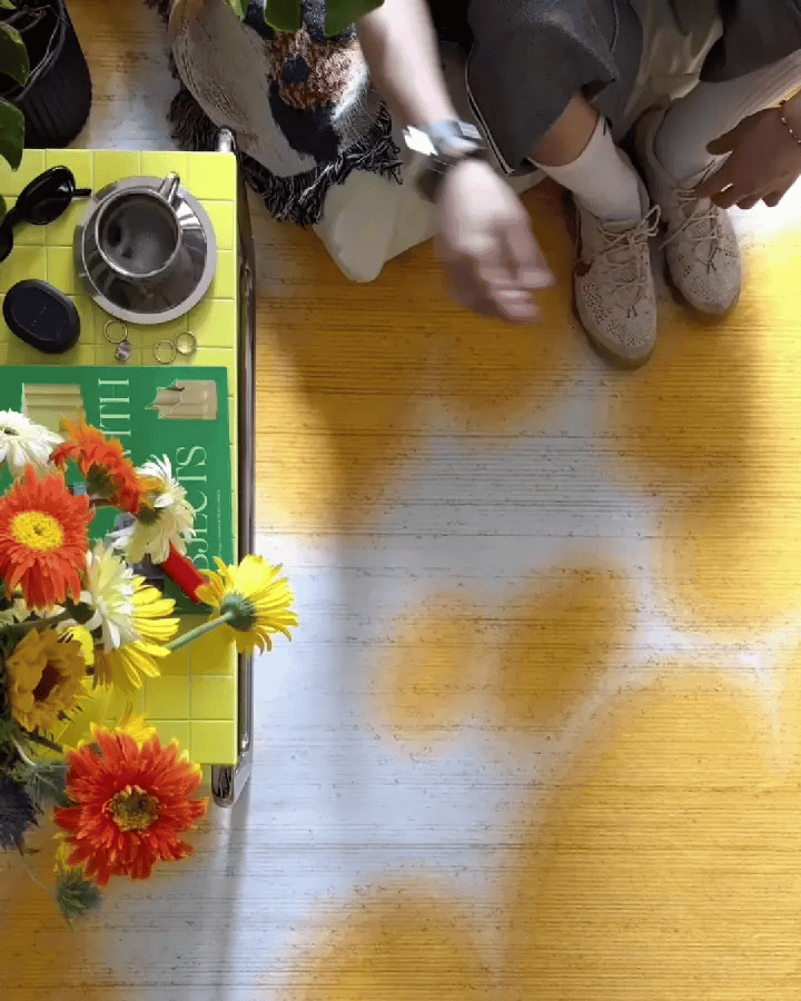 A man sitting on a white togo sofa resting his legs in beige fly knit sneakers on a yellow and off white floral cowhide area rug, with a yellow tiled coffee table, vibrant flowers, and colorful dopamine home decor in the living room.