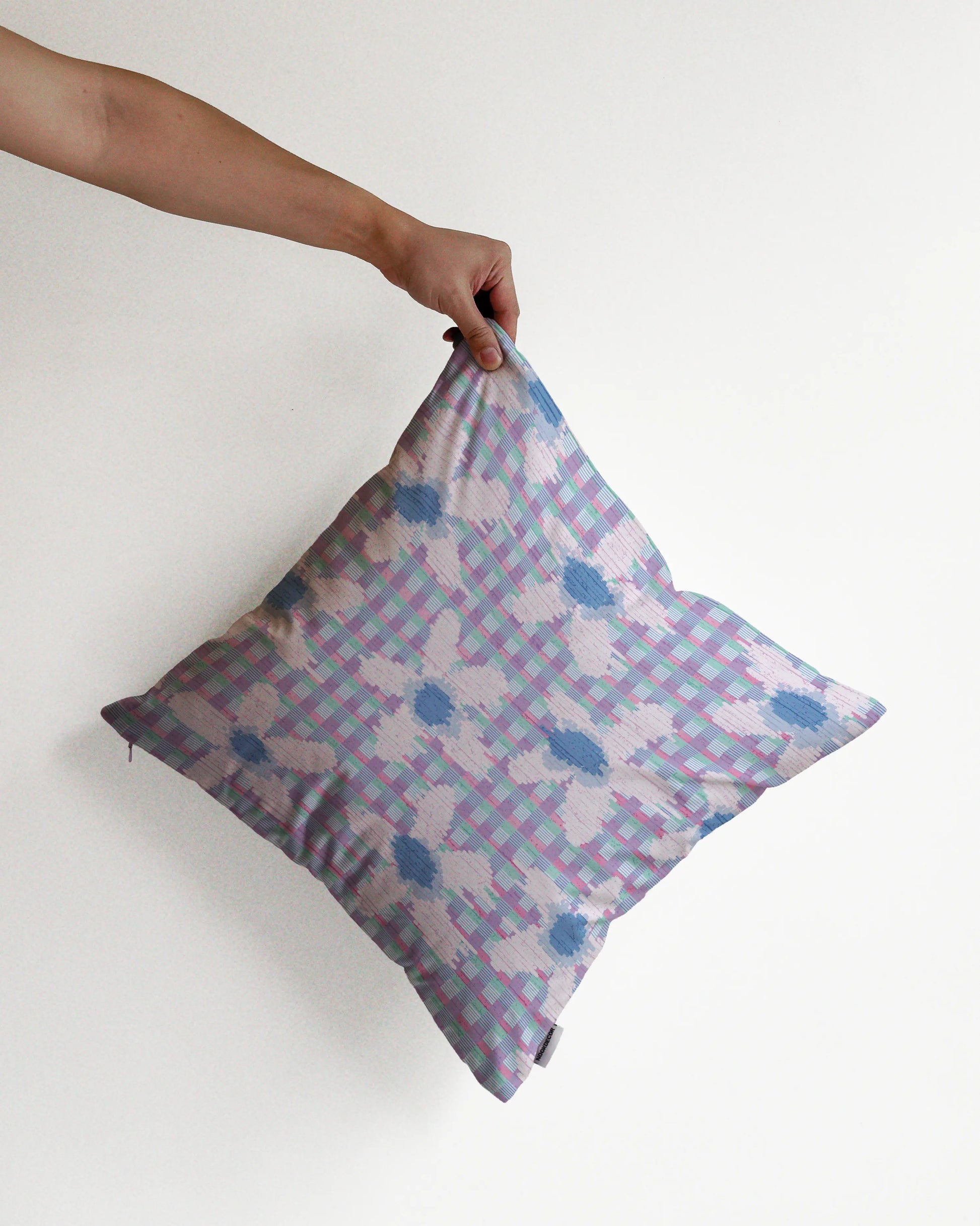 A hand holding an unique floral checkered cushion cover in pastel purple, pale blue, green hues, against a minimalist beige background, perfect for eclectic dopamine decor in a modern apartment.