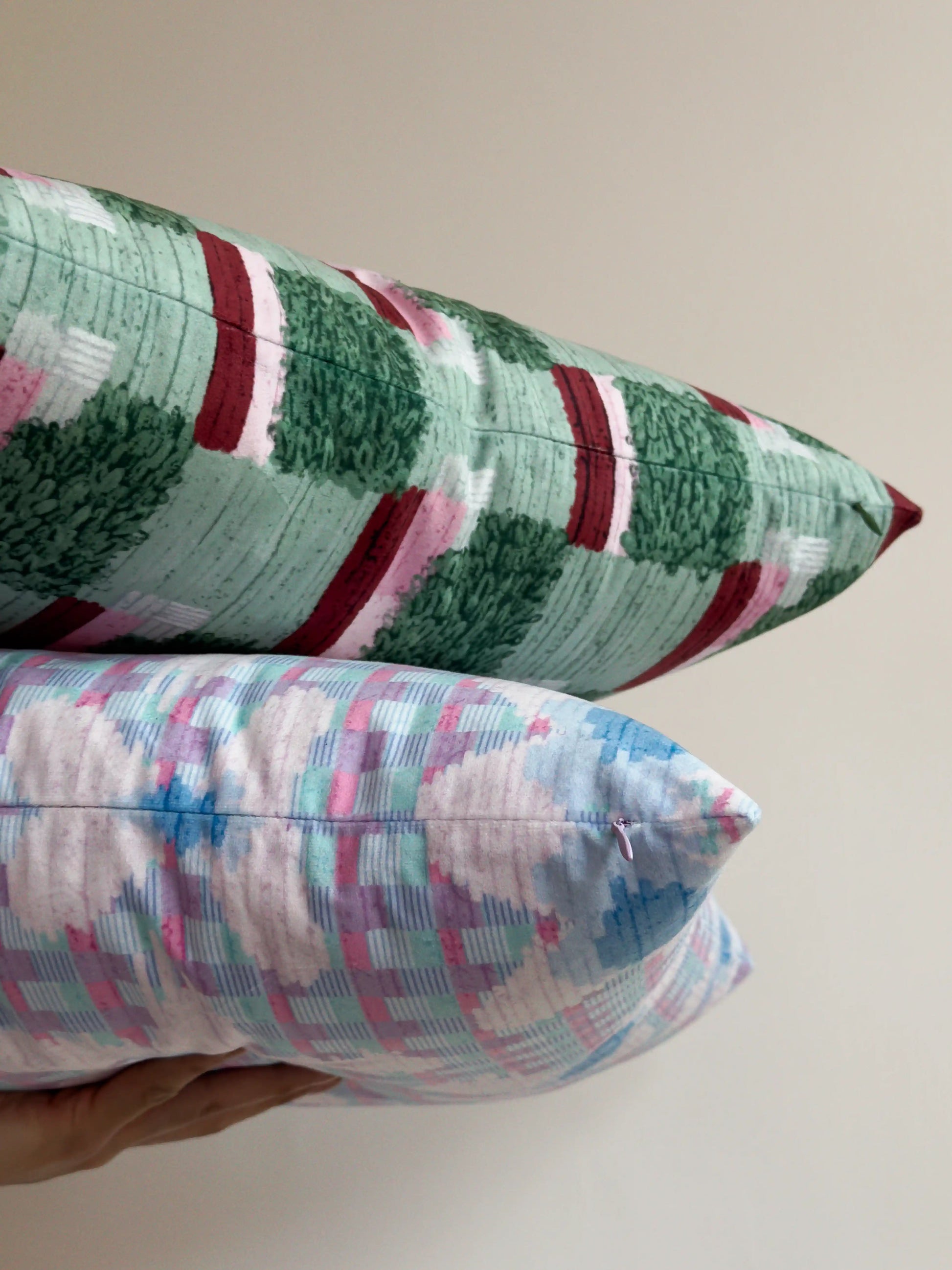 A dark green, light green and pink throw pillow and a pastel purple floral checkered throw pillow held against beige white wall, showing mix of dopamine, bold and maximalist home styling.