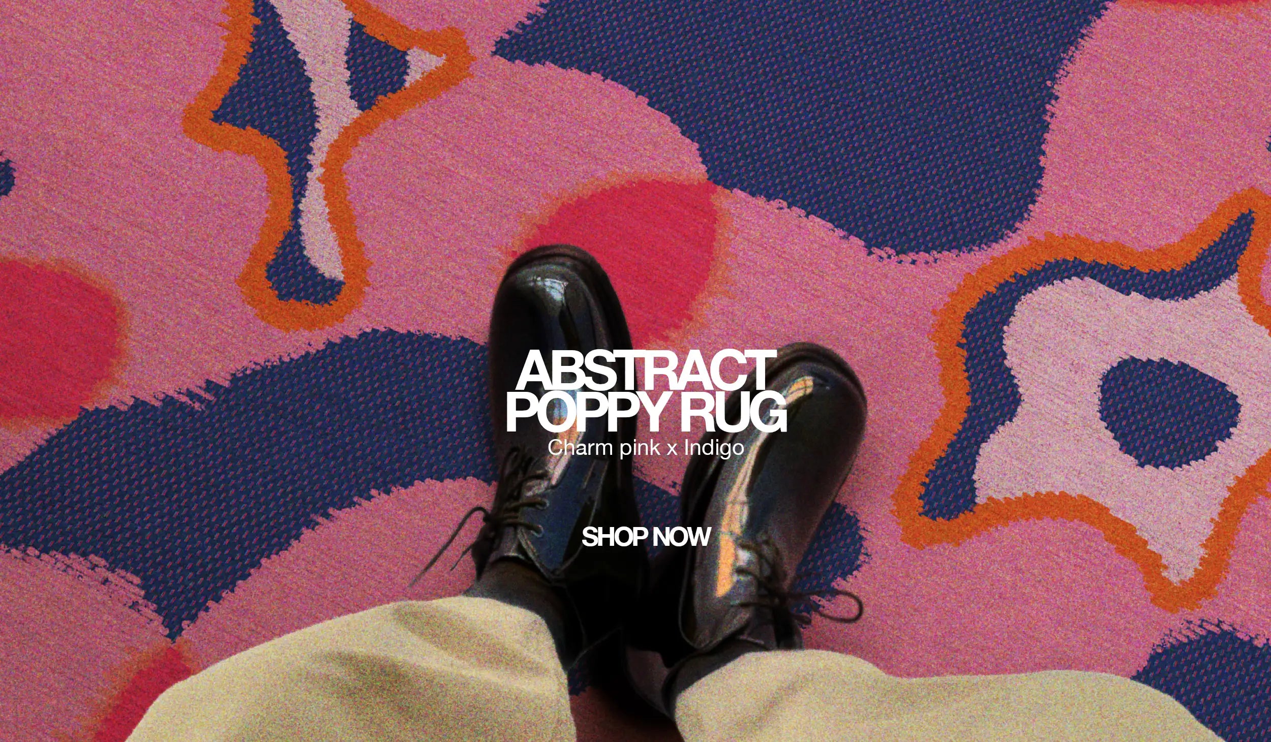Homepage banner, desktop version of a man wearing black leather shoes and beige pants is resting his leg on a large pink and dark blue floral area rug.
