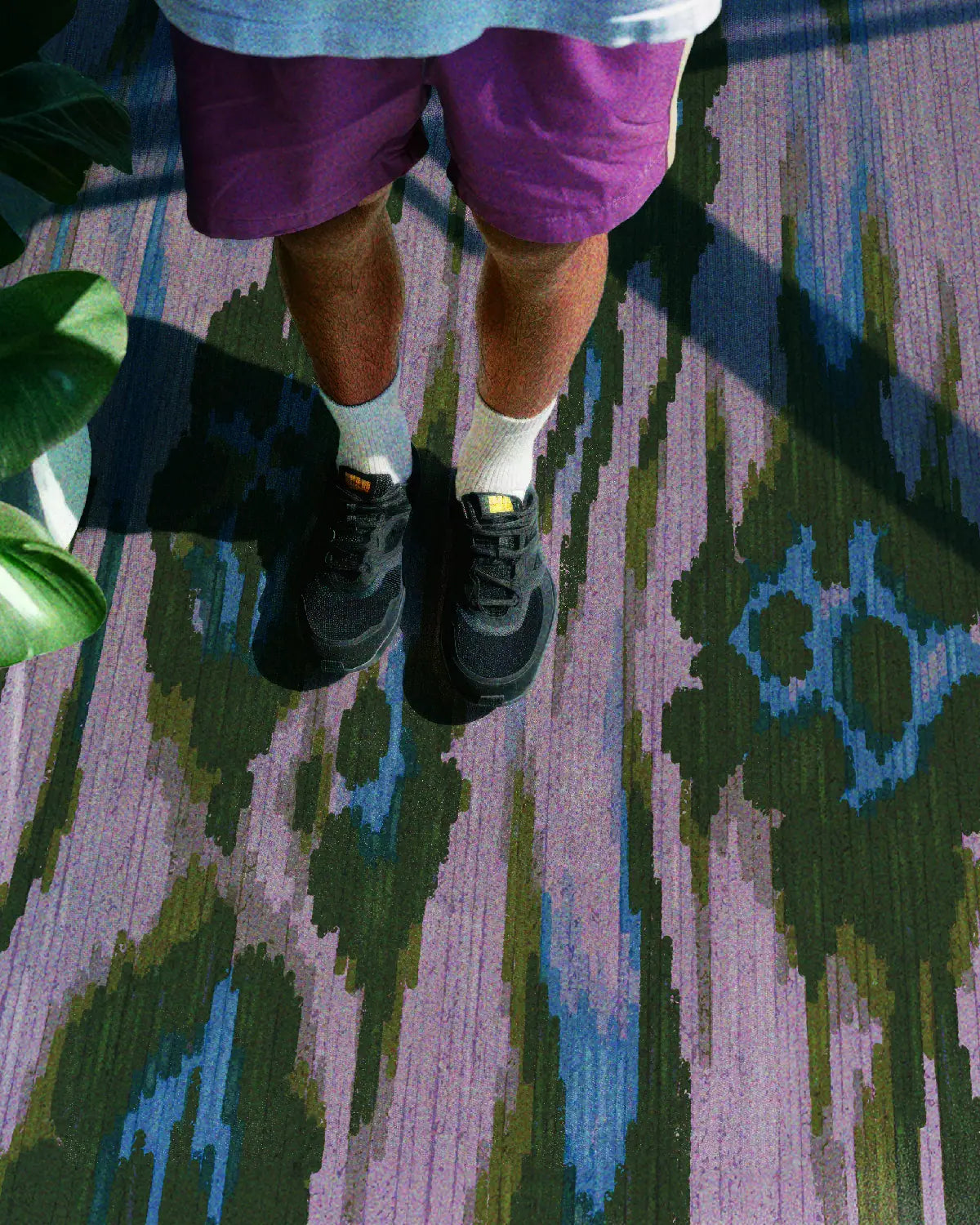 The man wears purple shorts and black sneakers, stands on a coquette rug; the colorful area rug features a leopard pattern in purple, blue, and green hues. Eclectic home decor for the living room.